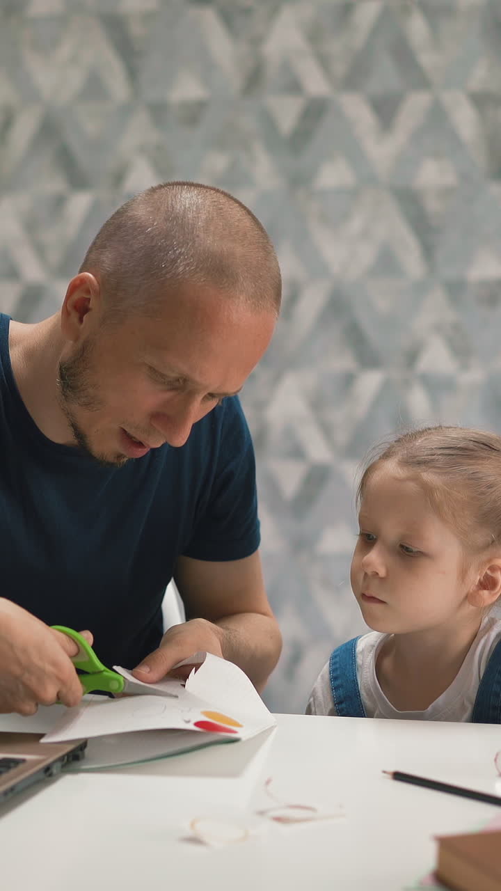 concept of distance learning young bald bearded dad cuts out figurine of paper in cage, shows little daughter student with blue eyes, she rejoices in result, they smile, laugh, sitting at table