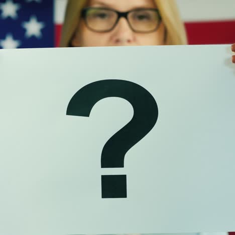 A-Woman-Holds-A-Poster-With-A-Question-Mark-Against-The-Background-Of-The-American-Flag