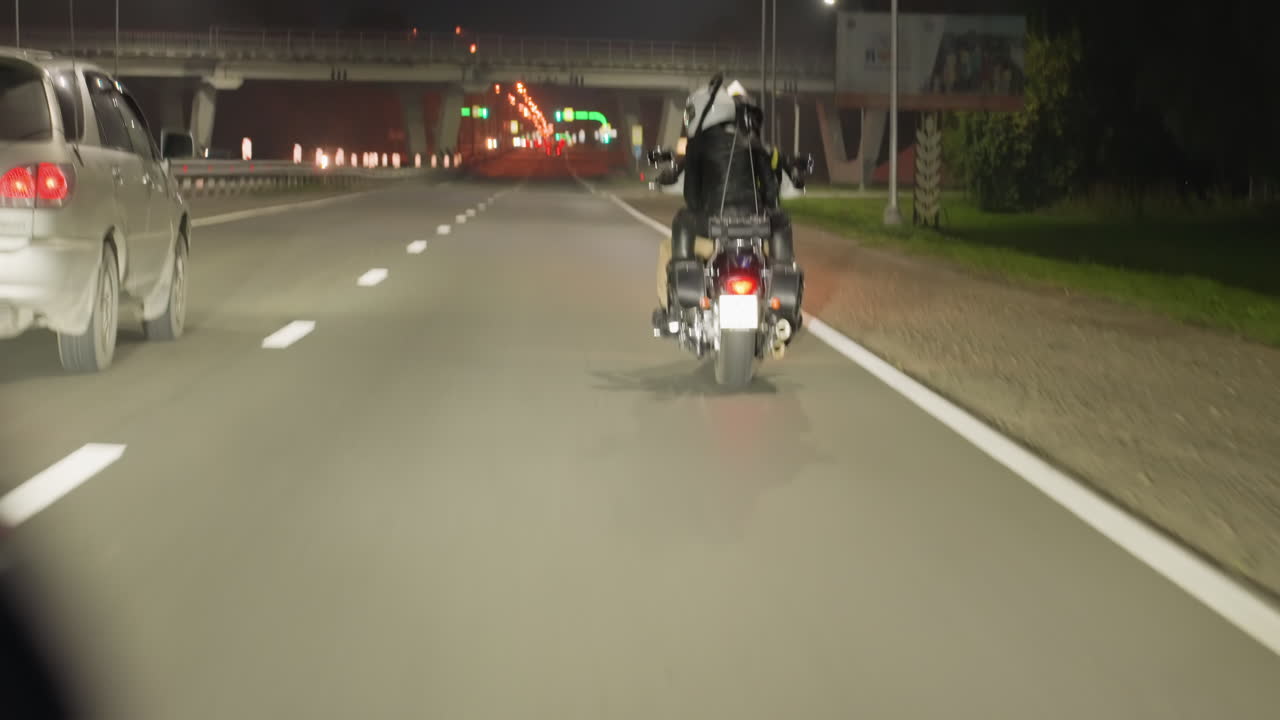Back view of motorcycle rider with passenger traveling highway at night, overtaking car approaching bridge ahead, streetlights illuminating road leading into city with glowing lights in distances