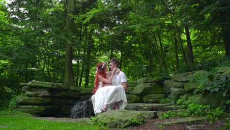 cheerful couple resting on stone near waterfall in forest. love couple kissing