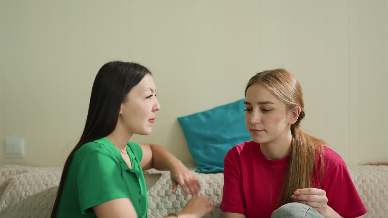 두 여성이 거실 소파에서 속삭이고 있어요. 한 명이 몸을 기울여 속삭이는 동안 다른 한 명은 이야기를 경청하며 비밀을 나누고 위로를 주고받는 모습이에요. 따뜻한 실내 조명, 편안한 옷차림, 차분하고 지지적인 분위기예요.