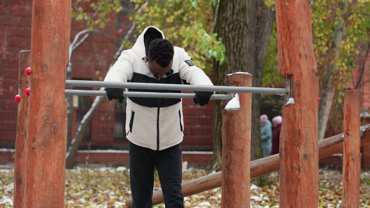 흑인 남자가 머리를 숙이고 앞에 철 막대기를 들고 휴식을 취하고, 따뜻한 털 재과 장갑을 입고, 겨울에 강렬한 운동에서 회복하고, 배경에서 일하는 사람들