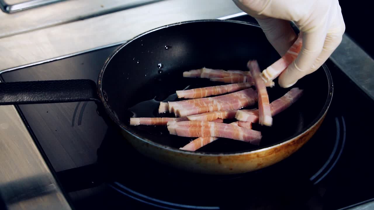 Frying bacon in the restaurant. Bacon pieces fry in a pan. Chef's hand in glove puts bacon cut into long strips on frying pan. Close-up