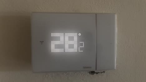 close-up of a digital thermostat displaying 28°c. camera tilts down from the wall to the device, concept of summer heat, climate control, and indoor temperature regulation
