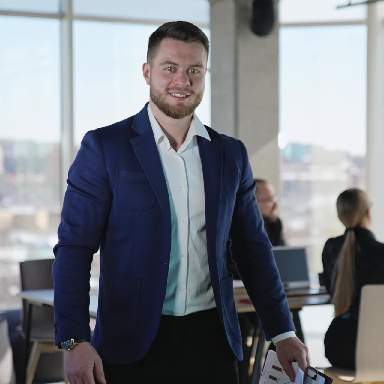 Middle-aged male businessman standing in the office and smiling to the camera. Teammates working at the table at the backdrop