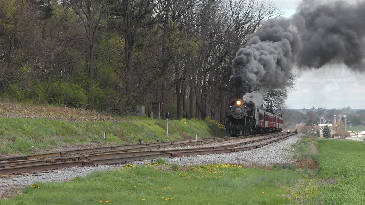 A steam locomotive chugs along scenic tracks, releasing puffs of smoke as it moves through lush green fields. The journey captures the charm of vintage trains in a beautiful rural landscape