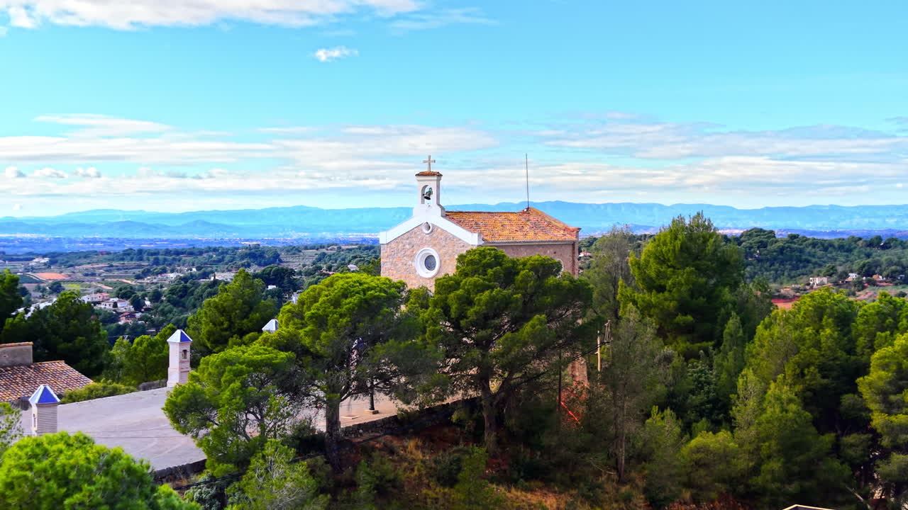 Edificio tradizionale di chiesa cristiana su un fianco di collina con vista su un vasto paesaggio e una catena montuosa sotto un cielo nuvoloso. Piccola cappella bianca con campanile tra i pini. Spagna.