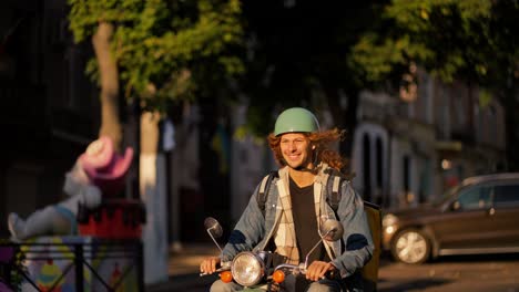 a guy with long curly hair in a denim jacket on a green moped rides along the road in a beautiful summer city