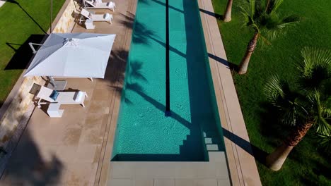 Aerial-View-of-a-Luxurious-Villa's-Pool-with-Palm-Trees-and-Sun-Umbrellas,-Surrounded-by-Lush-Lawn