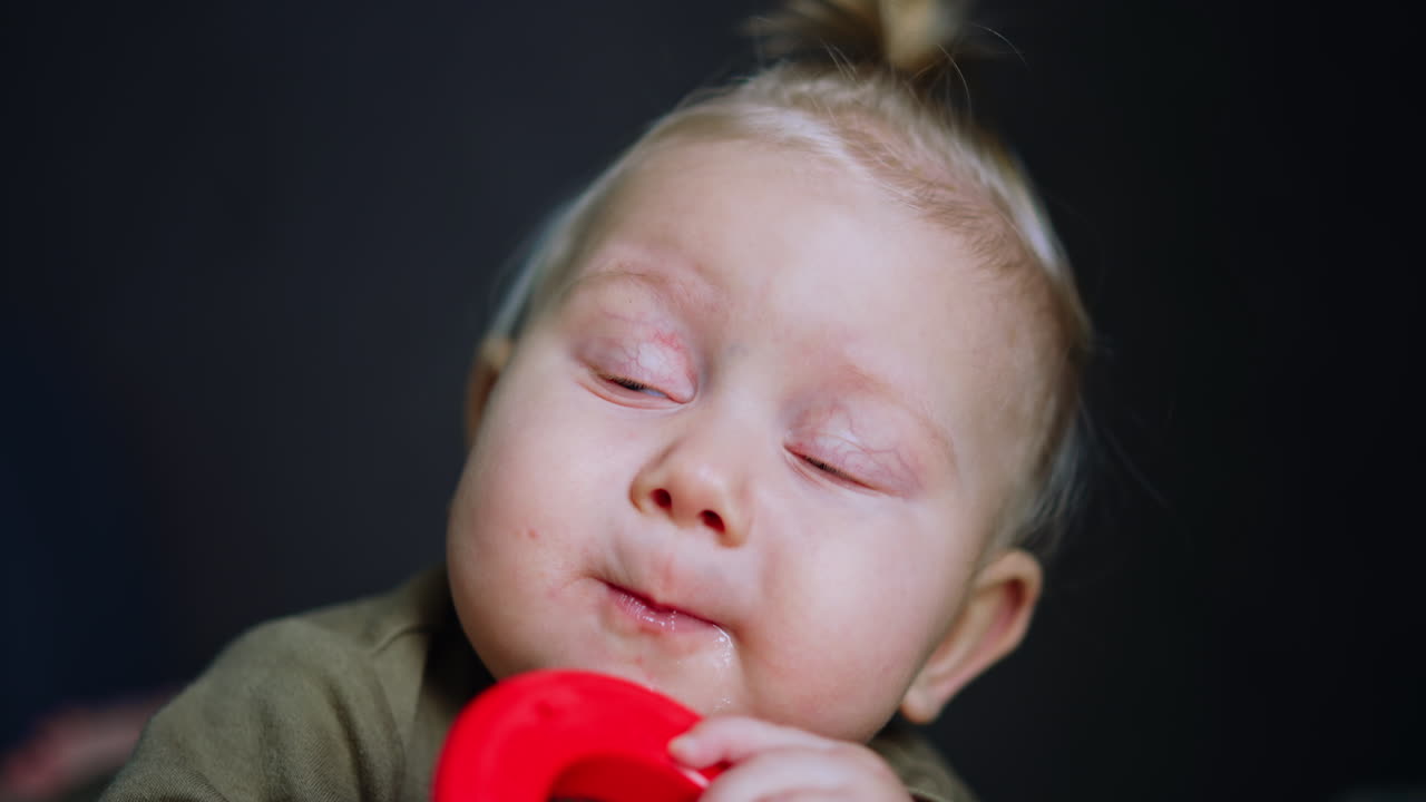 Lovely cute Caucasian baby with blue eyes and blond hair. Pretty child chewing red plastic ring. Close up.