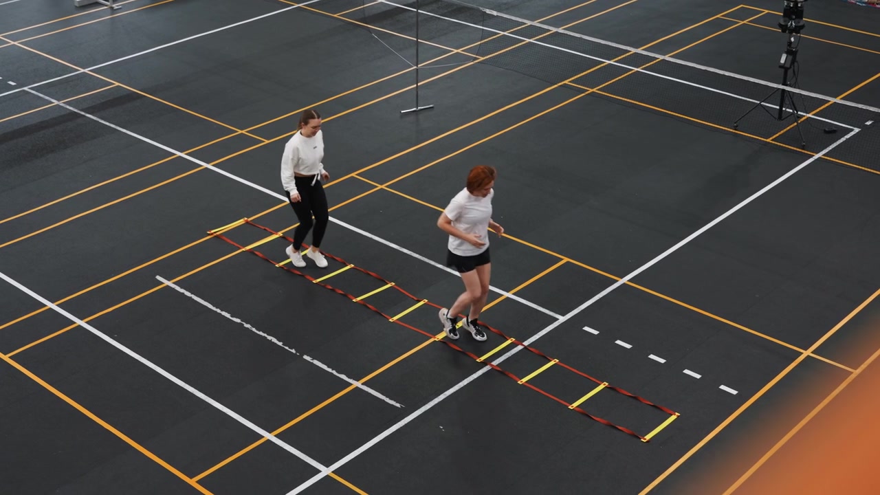 Allenatore e atleta su un campo da badminton che eseguono un esercizio di agilità con la scaletta. Intensa sessione di allenamento focalizzata in una palestra al coperto con linee segnate e pavimento nero. L'allenatore dimostra un passo.