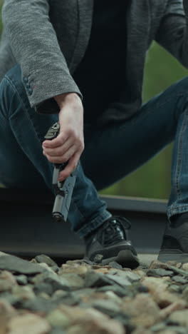 una vista cercana de una persona con una chaqueta gris, pantalones vaqueros azules y zapatos de lona negros sentados en una vía ferroviaria, sosteniendo una pistola con un dedo en el gatillo, vista borrosa de un bosque verde