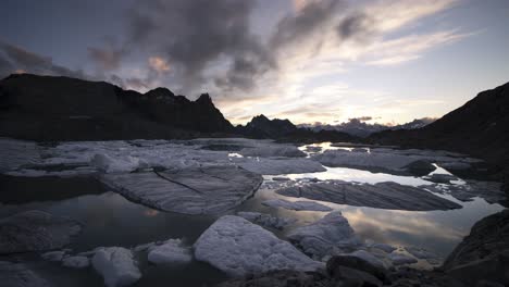 day to night sunset time-lapse of a lake full of icebergs from a melting glacier in the remote swiss alps as the sky changes from glowing to full of stars before being engulfed by clouds and darkness