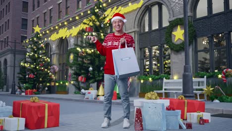 full body of asian man in santa suit with the shopping bags smiling and pointing to the side while standing in christmas night