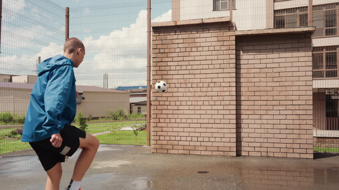 Hvit mann sparker ball i regnet, blå jakke, murvegg som mål, sølepyttsprut, urbant område, fokusert trening, solo-spiller som øver på returer og ballkontroll, våt betong og overskyet himmel