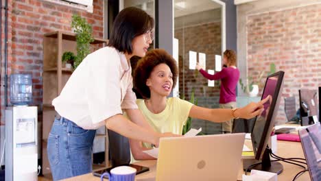 Two-happy-diverse-female-creative-colleagues-in-casual-discussion-at-desk-in-office,-slow-motion