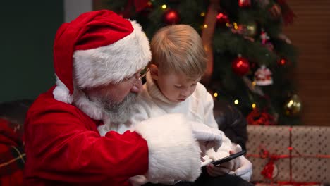 santa claus with a little boy clicking on a smartphone, choosing a christmas present on the internet. e commerce. concept of the new year spirit, holidays and celebrations. close up. slow motion