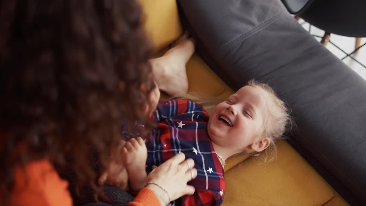 Loving mother tickling baby daughter little girl on sofa, sincerely laughing