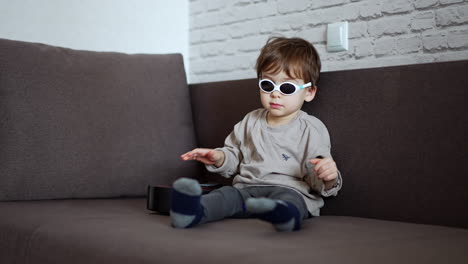 dark-haired toddler in sunglasses touches the strings of a guitar beside him. lovely kid plays the guitar and takes off his glasses.