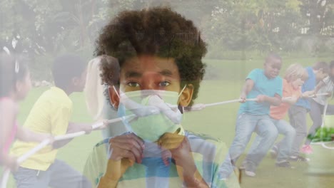African-american-boy-wearing-a-face-mask-against-group-of-diverse-kids-playing-tug-of-war-in-garden