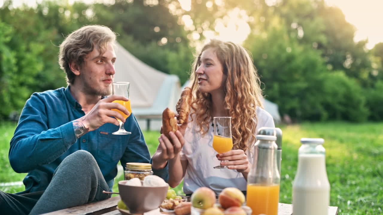A couple having breakfast in the nature at glamping. Drinks, food, talking and smiling. Tent on the background. Slow motion