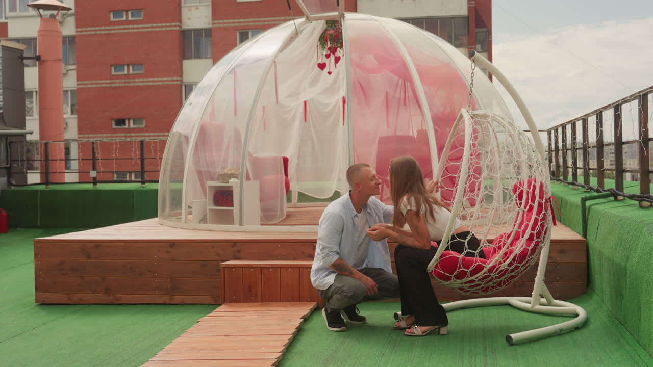 rooftop proposal outside clear bubble dome, man kneeling before partner on wooden platform, pink cushions inside transparent dome, artificial turf and urban skyline backdrop, string lights