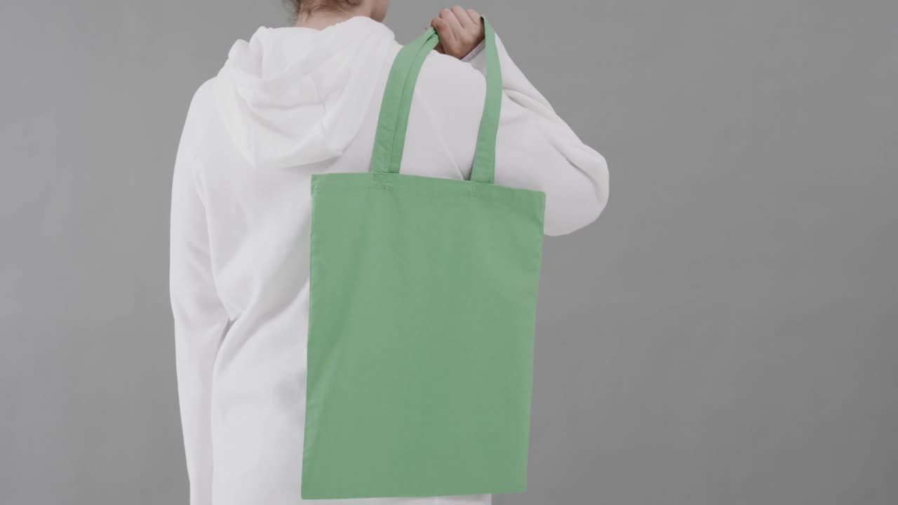 Donna caucasica che indossa una felpa bianca con cappuccio, tiene una borsa verde su sfondo grigio, spazio per il testo, movimento rallentato