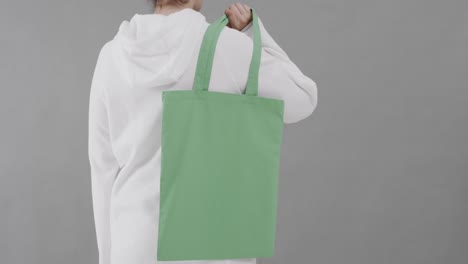 caucasian woman wearing white hoodie holding green bag on grey background, copy space, slow motion