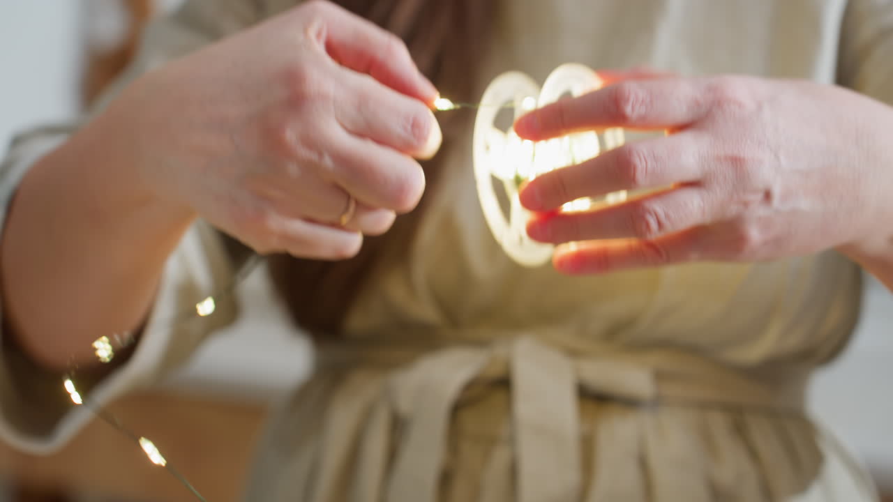 Close-up of woman's hand in brown gown carefully rolling out Christmas lights for decoration, preparing for festive holiday celebrations, setting up cozy Christmas atmosphere