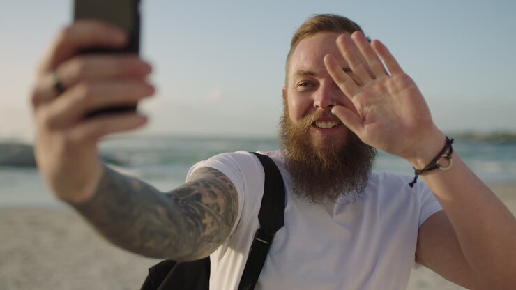 Flot hipster mand med skæg bruger telefon til videochat på stranden, taler og vinker.