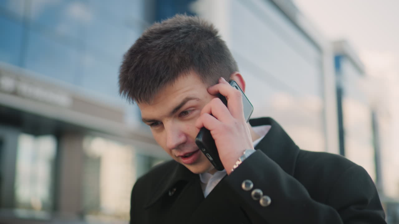 검은 코트를 입은 남자가 현대적인 유리 건물과 주차된 자동차가 있는 활기찬 도시 지역에서 의견 불일치로 머리를 흔들면서 두 개의 전화로 대화를 나누고 있습니다.