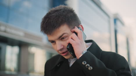 hombre con abrigo negro hablando en dos teléfonos indistintamente mientras sacude la cabeza en desacuerdo en una vibrante zona urbana con modernos edificios de vidrio, coches estacionados