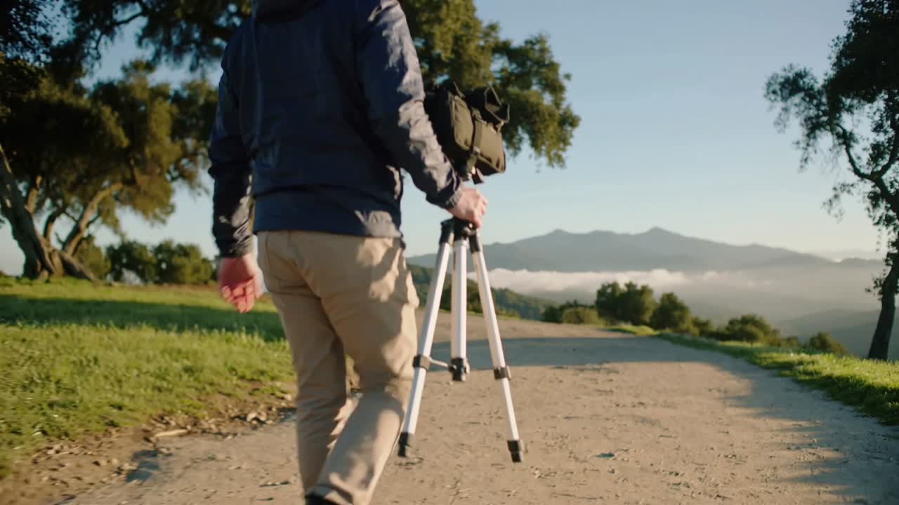 Walking navy-jacketed man carrying tripod and camera bag on hillside trail, moving to shoot valley