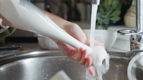 thorough washing of electric hand blender under running tap water by cook with visible traces of red berry on fingers while carefully rotating tool head to ensure proper cleaning