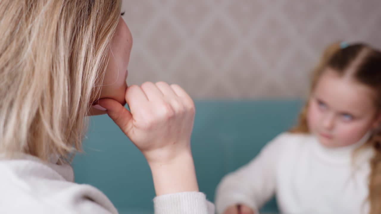 흰색 스웨터를 입고 포니테일을 한 금발 머리 소녀가 테이블에 앉아 엄마가 자신을 가리키는 것을 보며 집중하고 있어요. 이는 훈육, 양육, 어린 시절 행동, 관심, 교육을 상징해요.