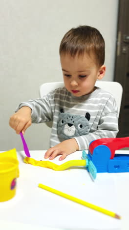 lovely baby sitting at desk uses a stick to cut the plasticine. toddler boy developing creativity doing art at home. vertical video.