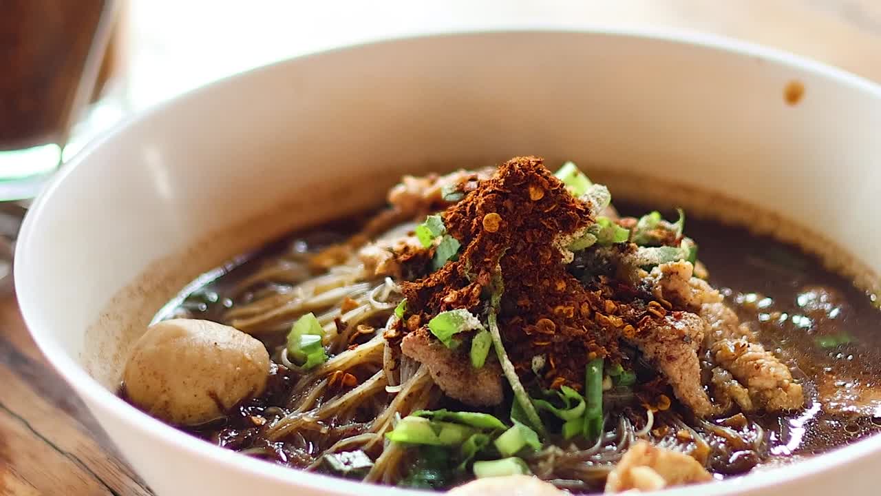 A close-up view of a noodle soup bowl topped with meat, vegetables, and chili flakes.