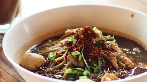 a close-up view of a noodle soup bowl topped with meat, vegetables, and chili flakes.