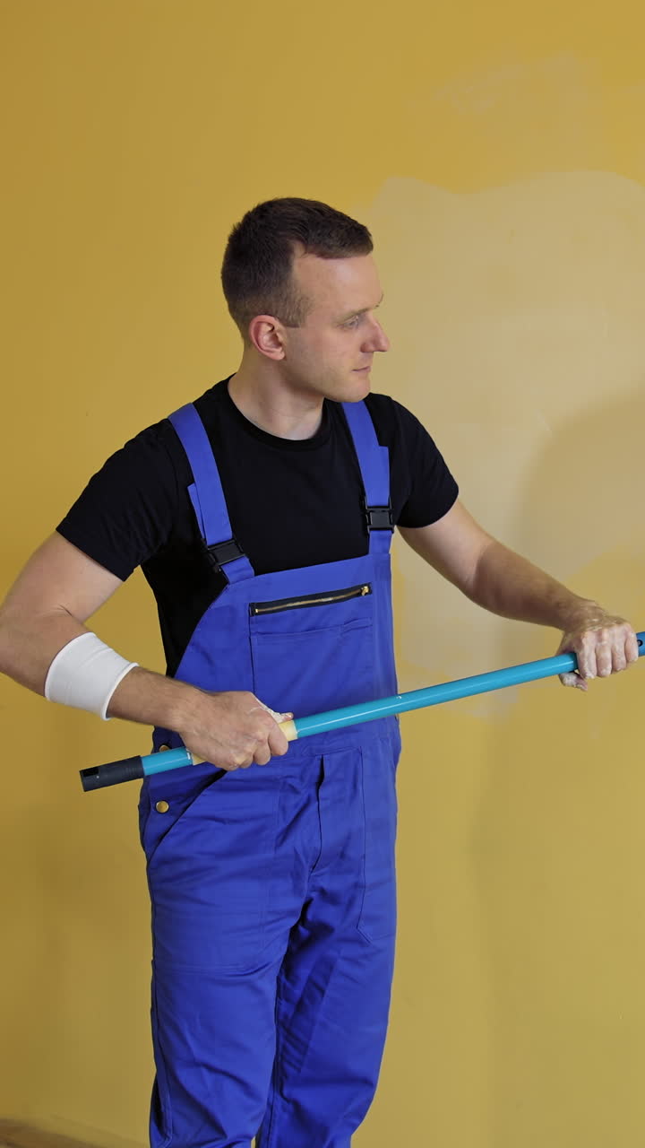 Man is painting walls at home. Young builder in blue uniform renovating walls with a paint roller on a long handle. Painting services. Vertical video