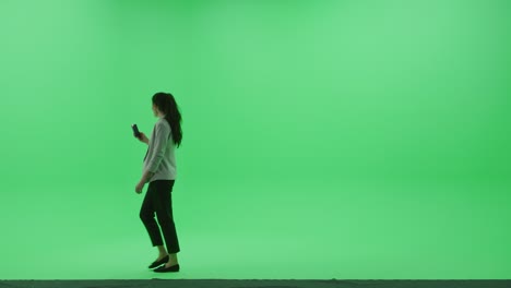green screen chroma key studio: beautiful mature woman wearing stylish business casual clothes uses smartphone to browse through internet and social media walks across the room. side view camera shot