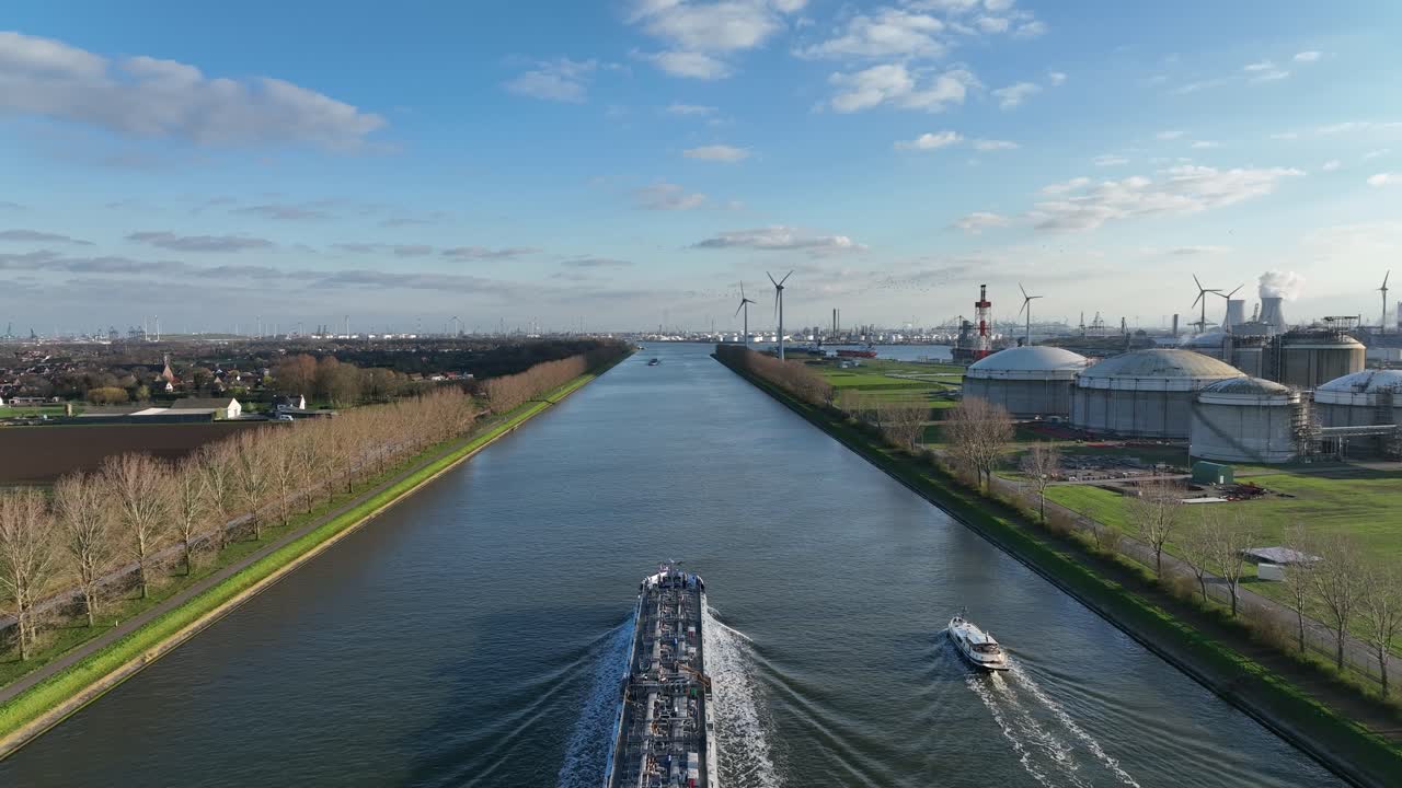 Zoom in aerial shot of cargo ship sailing Antwerp industrial canal surrounded by factories, wind turbines, and tanks on partly cloudy day