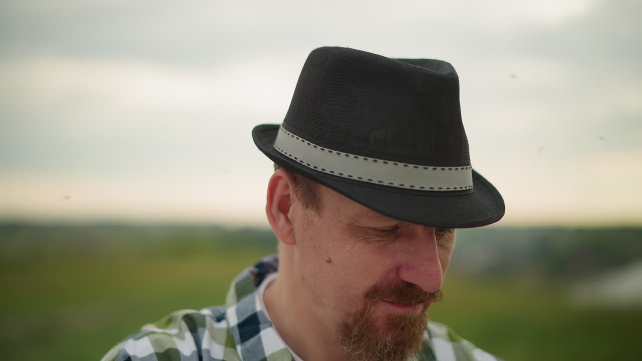 A man in a black hat and a checkered shirt is intensely focused on an unseen task, with mosquitoes visibly flying around his head. His deep concentration creates a serene yet determined atmosphere