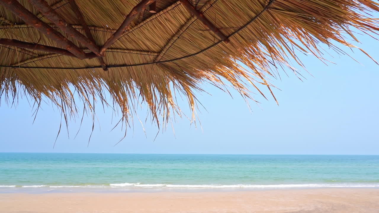 Tetto di fronde di palma offre ombra su un'ampia spiaggia sabbiosa con piccole onde al rallentatore che si infrangono sulla riva