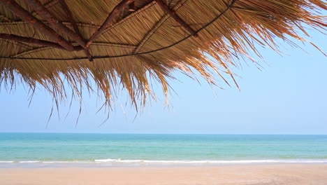 thatched roof made of palm leaves provides shade on wide sandy beach with slow motion little waves rolling onto the shore