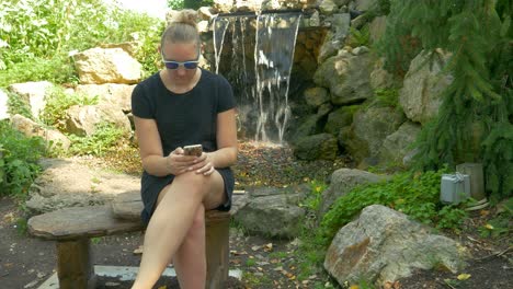 Una-Mujer-Alta-Con-Un-Vestido-Negro-Corto-Se-Sienta-En-Un-Banco-Cerca-De-Una-Cascada,-Inclinando-A-Una-Chica-Atractiva-En-Un-Parque-Usando-Su-Teléfono-Inteligente-Para-Enviar-Mensajes-De-Texto-A-Sus-Amigos