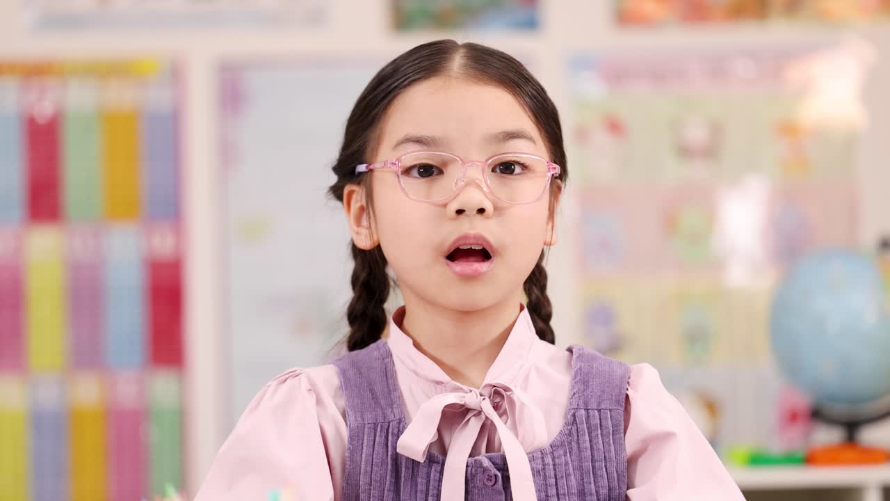 Elementary school girl with glasses expresses shock and surprise, hands raised, bright classroom lighting