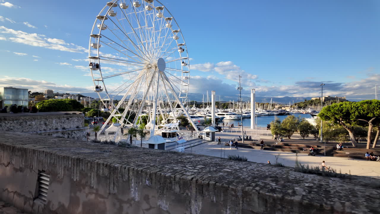 Side view of white ferris wheel rotating in Antibes, France with Port Vauban on the background