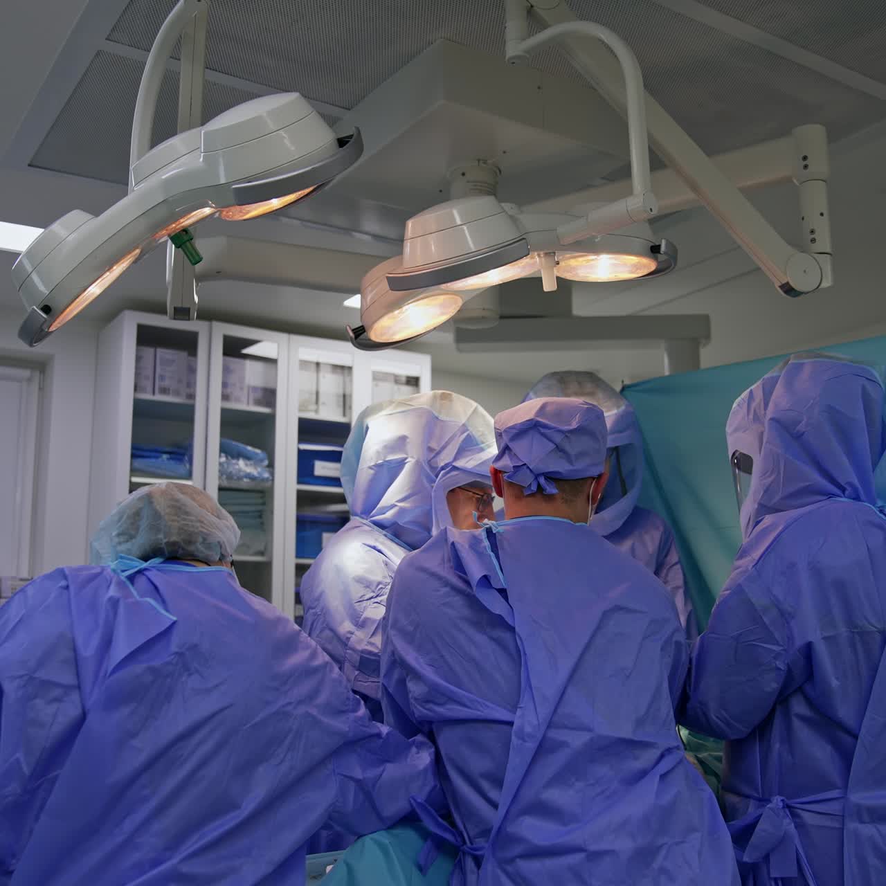 Medical team standing around the operational table under the bright lamps. Nurse passes the instruments to the surgeons
