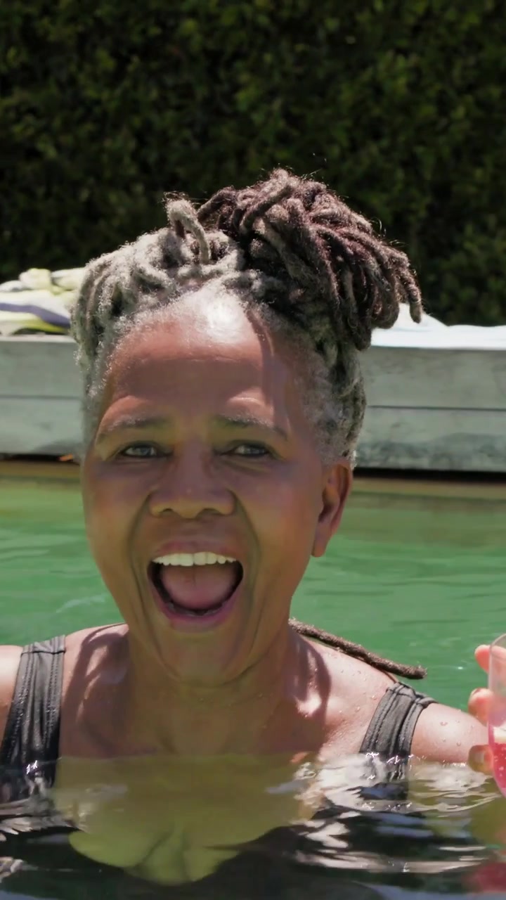 Vertical video: Laughing woman in swimsuit holding glass in home pool, friend arriving toasting