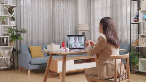 sick asian businesswoman holding a thermometer to the video call with doctor on a laptop while working at home